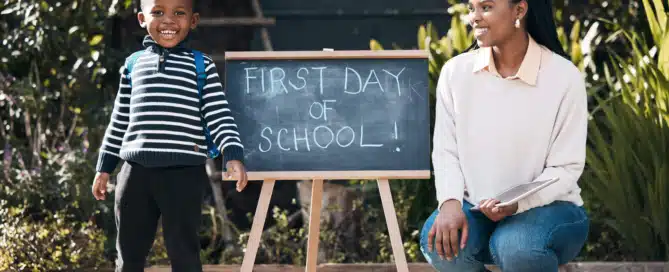 young boy smiling with backpack on standing next to sign that reads "First Day od School" with his mom kneeled down on the other side smiling